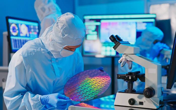 Engineer in a cleanroom suit examining a rainbow-coloured processed wafer